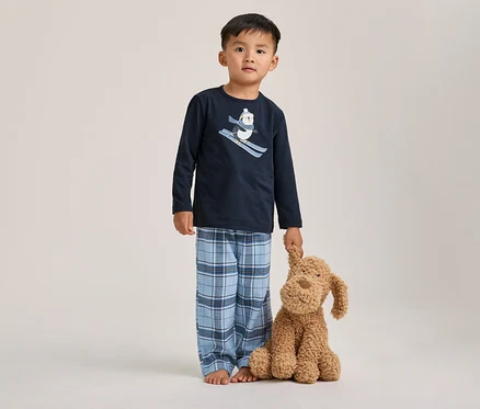 Ein kleiner Junge steht barfuß in einem dunkelblauen Pyjama mit einem Ski fahrenden Pinguin-Motiv und einer blau karierten Hose, einen braunen Stoffhund haltend.