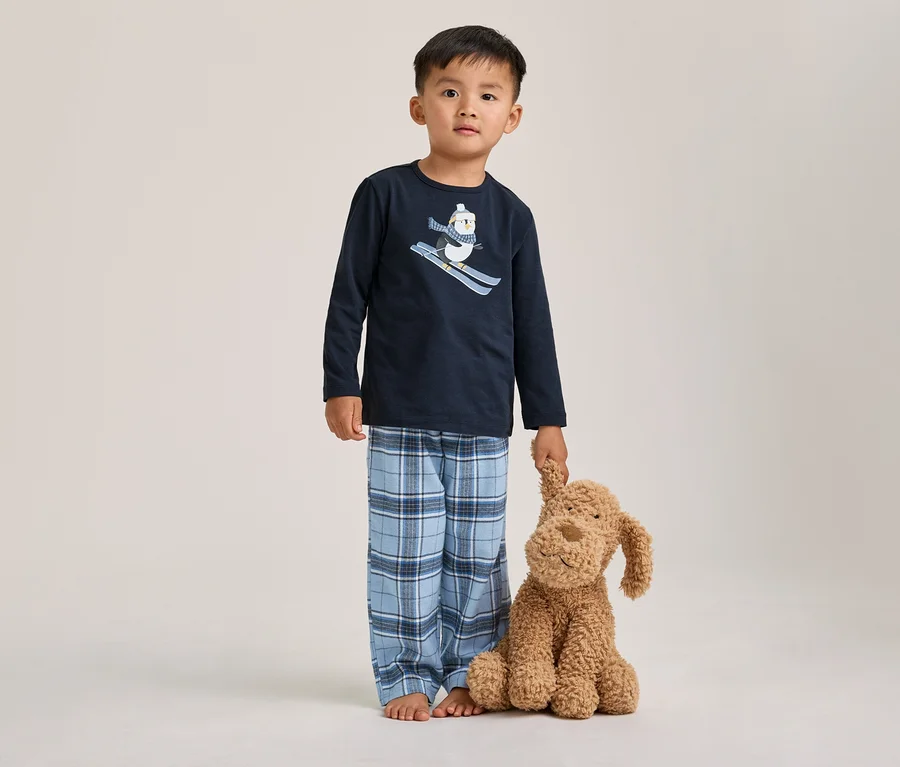 Ein kleiner Junge steht barfuß in einem dunkelblauen Pyjama mit einem Ski fahrenden Pinguin-Motiv und einer blau karierten Hose, einen braunen Stoffhund haltend.