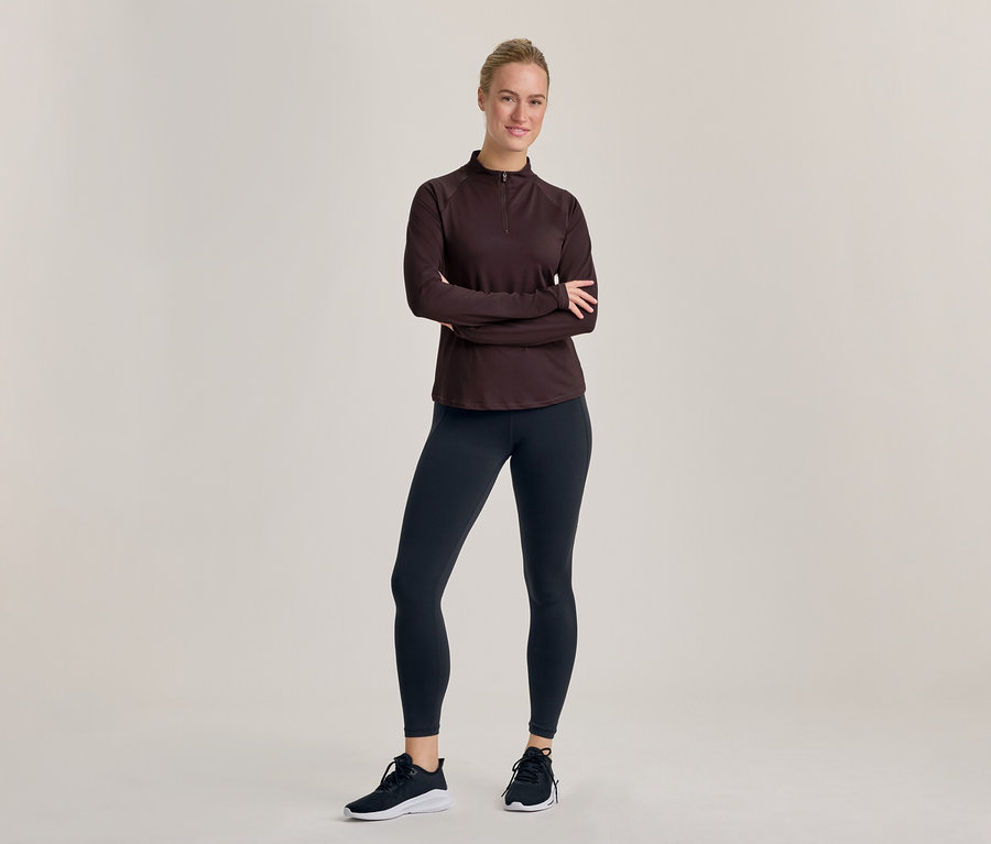 Eine lächelnde Frau steht mit verschränkten Armen, trägt ein dunkles langärmeliges Halb-Zip-Oberteil, dunkle Leggings und schwarz-weiße Sportschuhe.
