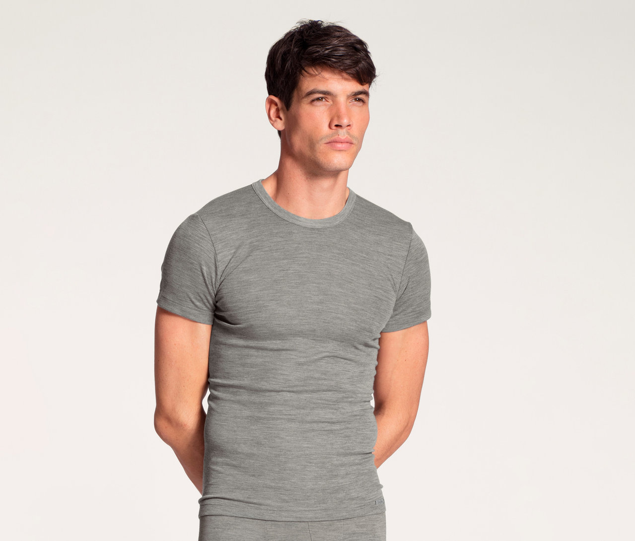 Mann in grauem Kurzarm-T-Shirt und passender grauer langer Unterhose, steht mit Händen hinter dem Rücken und blickt nach rechts.
