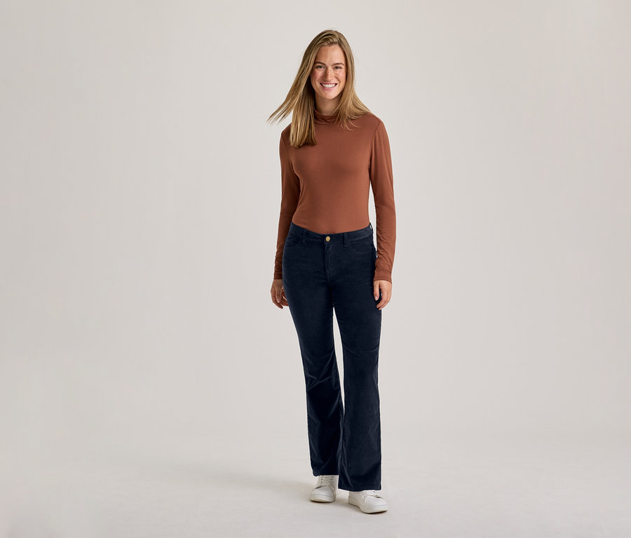 Eine lächelnde Frau steht in einem braunen Langarmshirt, dunkelblauen Cordhosen und weißen Turnschuhen.