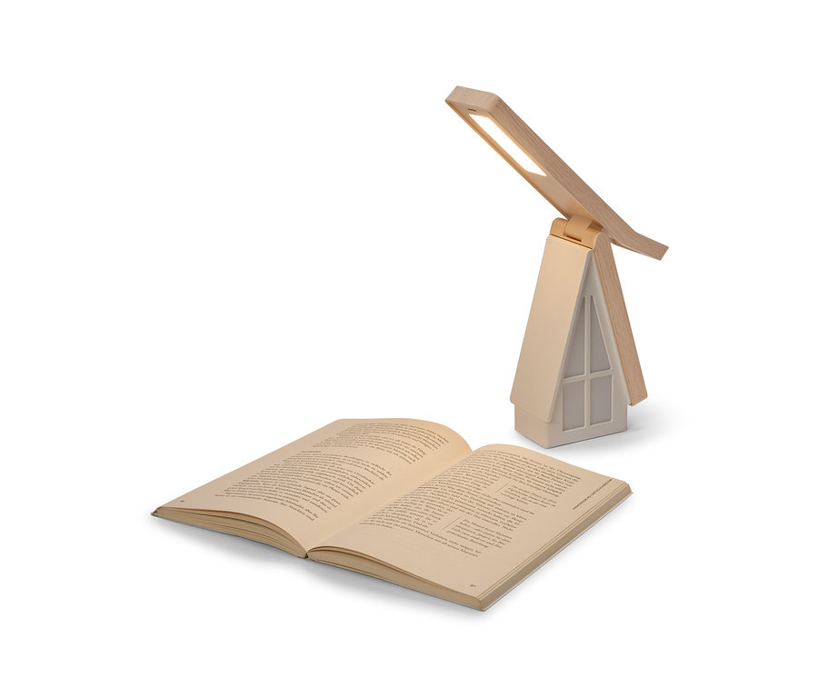 Ein aufgeschlagenes Buch liegt vor einer hausförmigen Tischlampe mit beigem Sockel und Holz-Dach. Die Lampe ist eingeschaltet und beleuchtet das Buch mit ihrem LED-Licht.