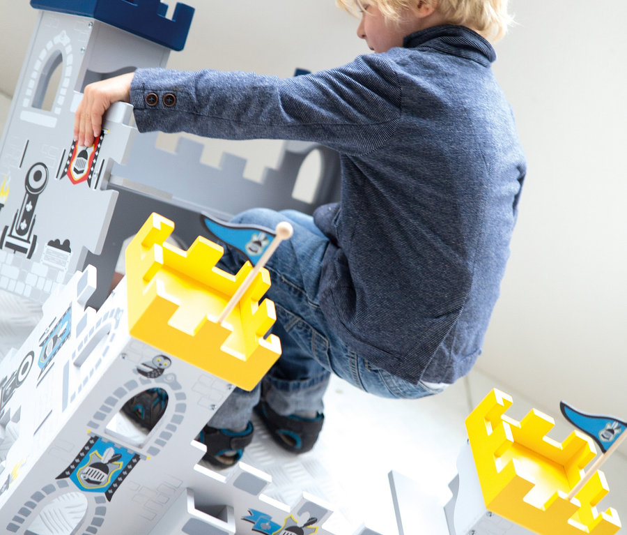 Ein kleiner blonder Junge in blauer Jacke und Jeans spielt mit einer großen grau-blauen Spielzeugburg, wobei er sich darüberbeugt und hineingreift. Die Burg hat gelbe Türme und blaue Flaggen.