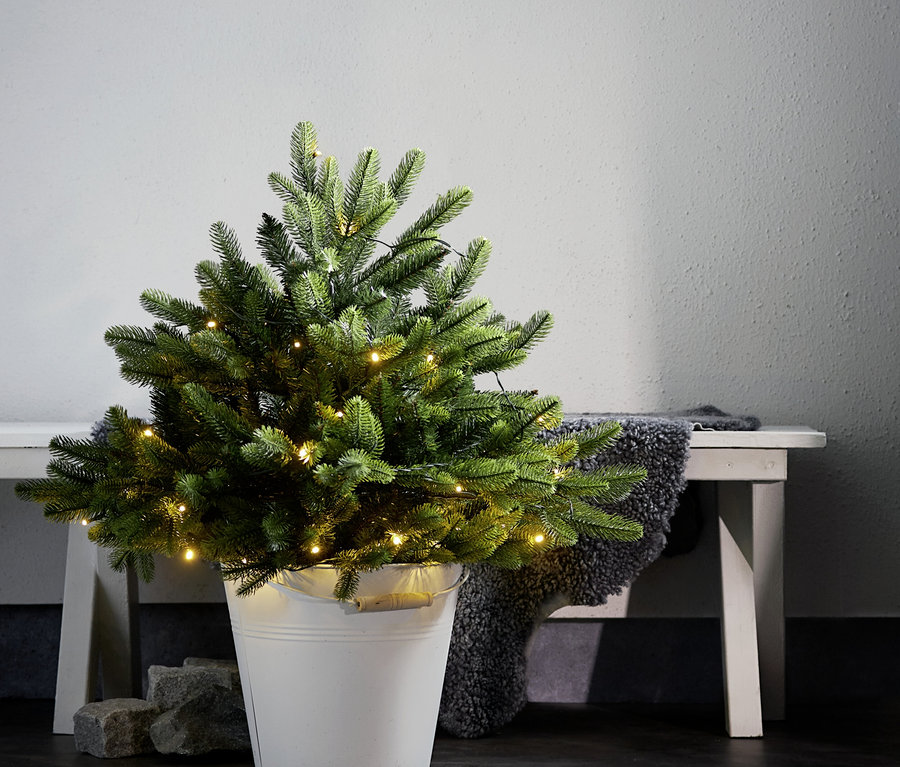 Kleiner künstlicher Weihnachtsbaum in einem weißen Eimer, mit warmen Lichterketten geschmückt. Dahinter steht eine helle Holzbank mit einem dunkelgrauen flauschigen Überwurf, daneben liegen zwei graue Steine am Boden.
