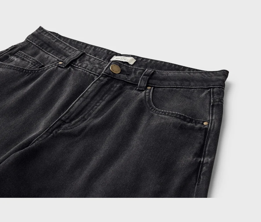 Nahaufnahme einer schwarzen Jeans aus gewaschenem Denim. Die Jeans hat einen Bund mit Gürtelschlaufen, einen goldfarbenen Knopfverschluss und Vordertaschen, darunter eine kleine Münztasche. Ein weißes Etikett mit „MY STYLE“ ist nahe dem Knopf sichtbar.