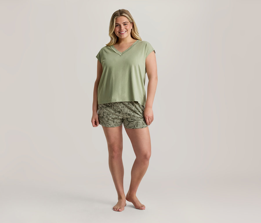 Lächelnde Frau in einem hellgrünen V-Ausschnitt-Oberteil mit Spitze und olivgrünen Shorts mit Blumenmuster, barfuß stehend.
