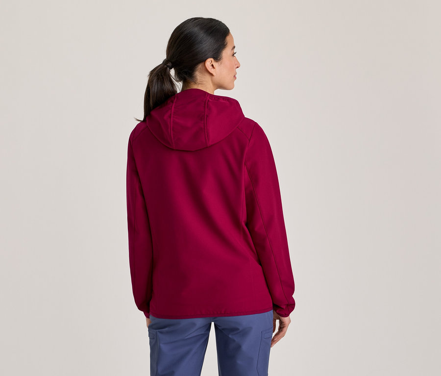 Frau von hinten gesehen, trägt eine magentafarbene Kapuzenjacke und blaue Hose, die Haare zum Pferdeschwanz gebunden, blickt nach rechts.