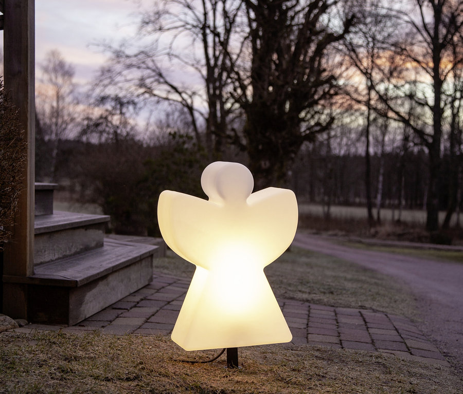 Leuchtende, engelförmige Gartenlampe steht im Gras neben einer Steintreppe und einem gepflasterten Weg in der Dämmerung.