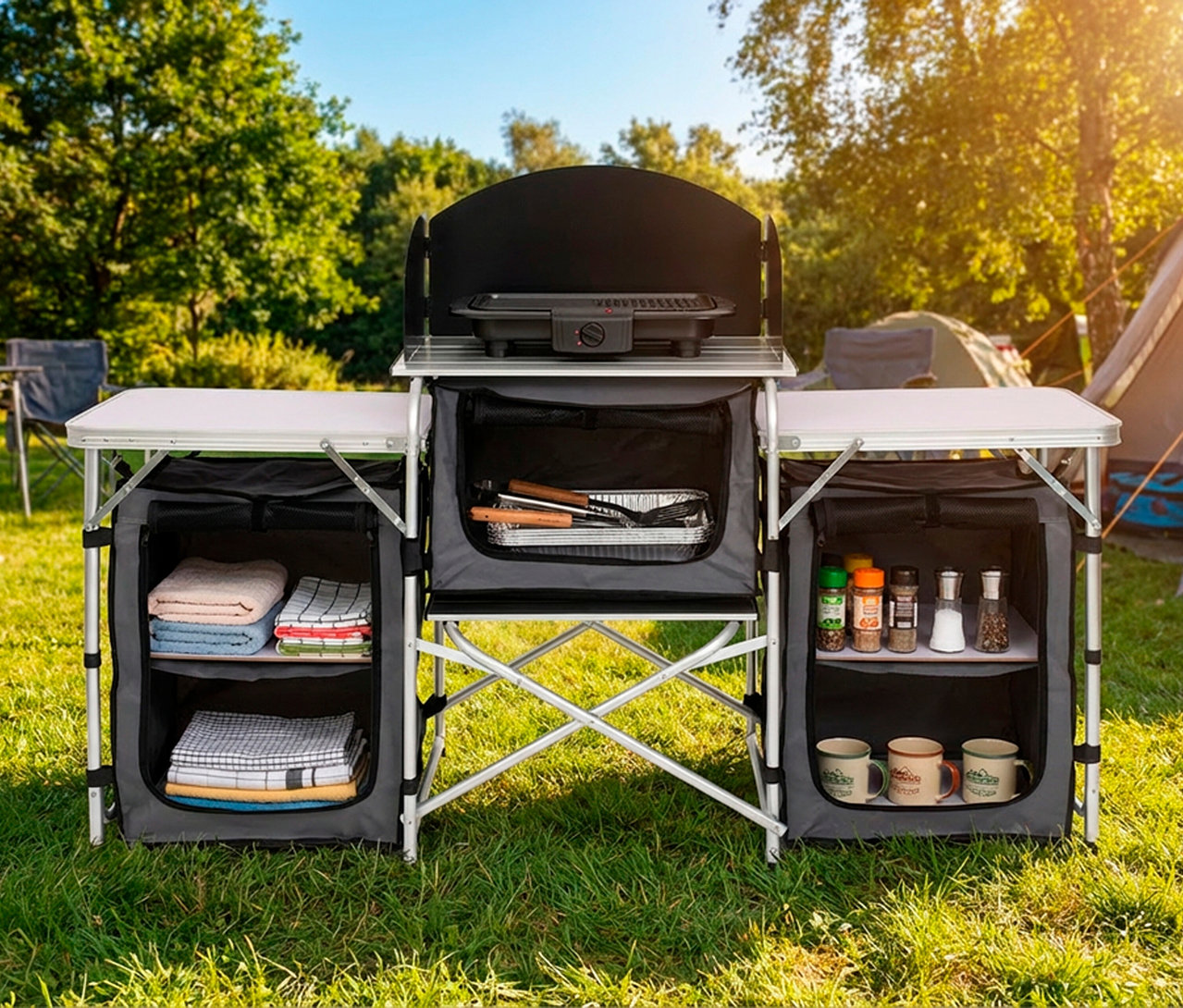 Faltbare Campingküche »Redwood M« auf grünem Gras. Sie hat eine zentrale Einheit mit Elektrogrill und Windschutz, flankiert von zwei Beistelltischen. Der linke Schrank enthält Handtücher, der rechte Gewürze und Tassen. Kochutensilien sind im mittleren Fach. Im Hintergrund sind ein Zelt und Bäume zu sehen.