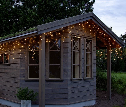 Holzgartenhaus mit warmweißer Eiszapfen-Lichterkette an der Dachtraufe. Daneben steht eine kleine Topfpflanze. Im Hintergrund sind Bäume und ein bewölkter Himmel zu sehen.