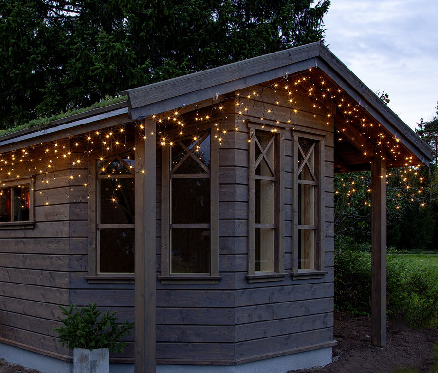 Holzgartenhaus mit warmweißer Eiszapfen-Lichterkette an der Dachtraufe. Daneben steht eine kleine Topfpflanze. Im Hintergrund sind Bäume und ein bewölkter Himmel zu sehen.