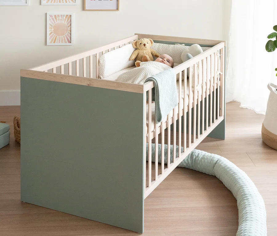 Ein Baby liegt in einem grün-hellholzfarbenen Gitterbett mit weißen Stäben, zugedeckt mit einer hellblauen Decke, neben einem Teddybär. Ein langer, hellblauer Zugluftstopper liegt auf dem Boden.