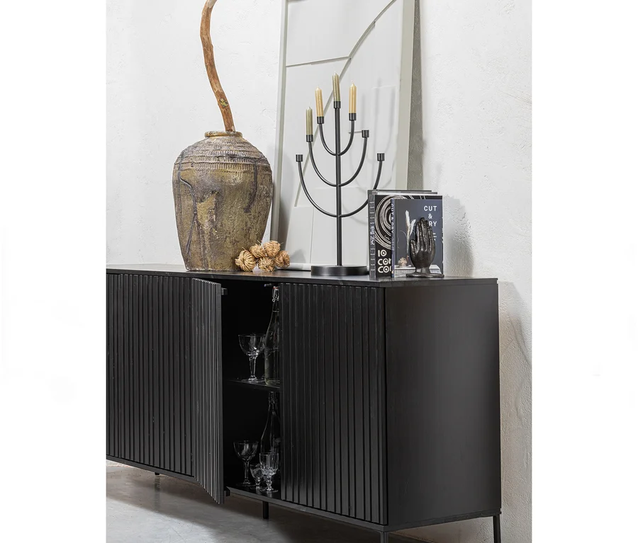 Schwarzes Sideboard mit geriffelten Türen, eine offenbart Gläser, darauf eine große strukturierte Vase mit Ast, ein schwarzer Kerzenständer mit Kerzen und Bücher.