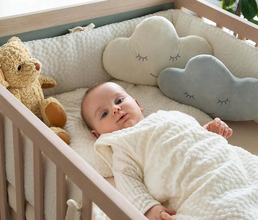 Ein Baby liegt in einem Holzbettchen, bedeckt von einem weißen Schlafsack, mit einem Teddybären und Wolkenkissen.