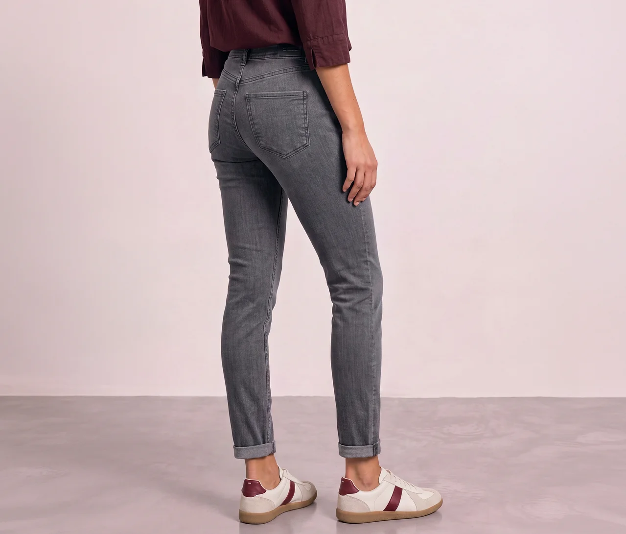Eine Person, von hinten gesehen, trägt eine dunkelrote Bluse, graue Street One Jeans mit umgeschlagenen Säumen und weiße Sneaker mit weinroten Streifen. Sie steht vor einer hellrosa Wand auf einem grauen Boden.