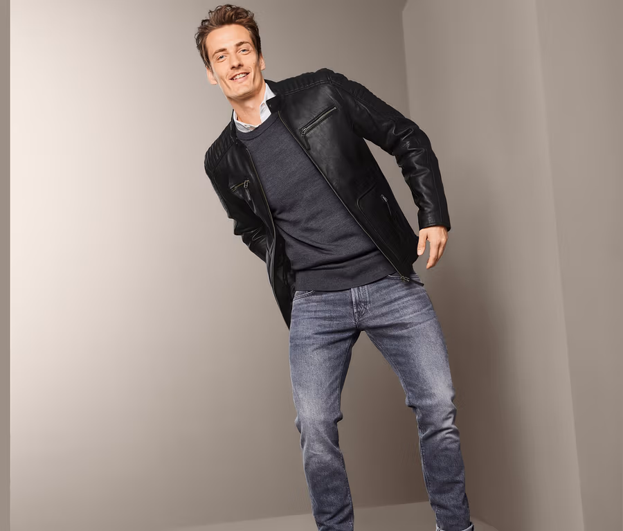 Ein Mann posiert in einer Lederjacke im Biker-Style, einem Pullover und Jeans.