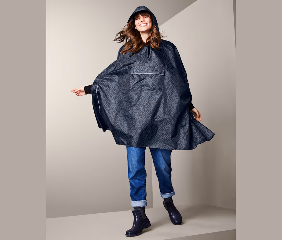 Lächelnde Frau posiert in blauem Regenponcho mit Kapuze, Jeans und Stiefeln.