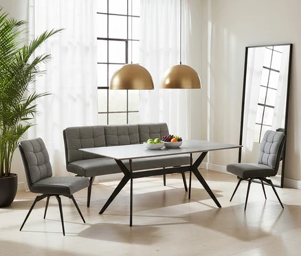 Modernes Esszimmer mit einem rechteckigen Tisch mit grauer Platte und schwarzen Kreuzbeinen. Eine graue, gesteppte Sitzbank und zwei passende Stühle umgeben den Tisch. Zwei goldene Hängelampen leuchten von oben, während auf dem Tisch Obstschalen stehen. Ein großer Spiegel und eine Pflanze schmücken den Raum.