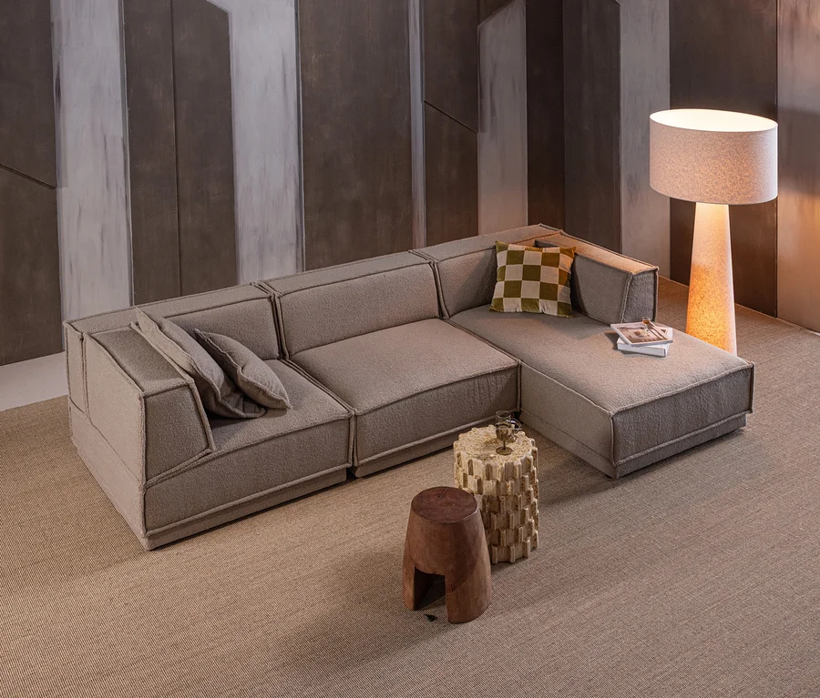 Ein modernes Wohnzimmer mit einem L-förmigen modularen Sofa aus hellbraunem Strukturgewebe, mit Kissen und einem karierten grünen Polster. Davor stehen ein dunkler Holzschemel und ein heller, zylindrischer Beistelltisch mit einem Getränk. Eine hohe Stehlampe mit Stoffschirm steht hinter dem Sofa, alles auf einem hellbraunen Teppich vor einer dunklen geometrischen Wand.