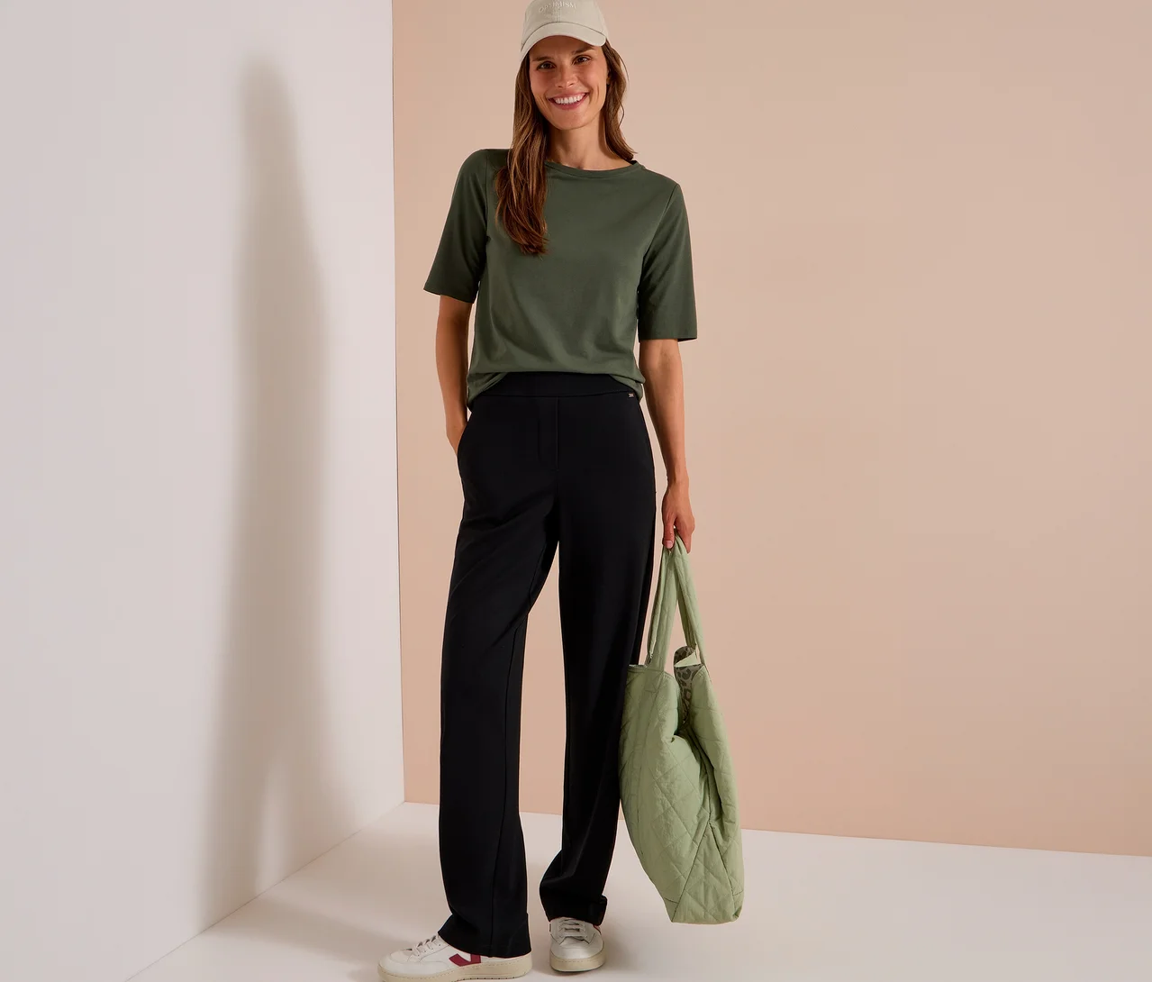 Eine lächelnde Frau trägt ein olivgrünes T-Shirt, eine schwarze CECIL Stoffhose, weiße Sneaker und eine beige Kappe, während sie eine gesteppte hellgrüne Tasche hält.