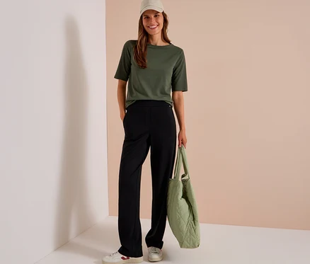 Eine lächelnde Frau trägt ein olivgrünes T-Shirt, eine schwarze CECIL Stoffhose, weiße Sneaker und eine beige Kappe, während sie eine gesteppte hellgrüne Tasche hält.