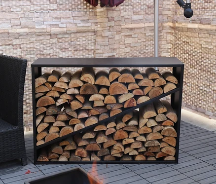 Ein ca. 114 cm großes Kaminholzregal aus schwarzem Metall ist mit Holzscheiten gefüllt. Im Hintergrund befindet sich ein Korbsessel.