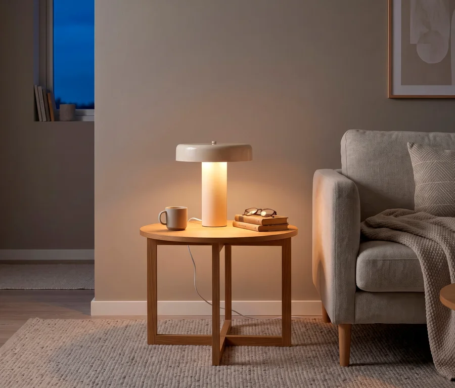 Eine cremefarbene Reality Tischleuchte "Erika" mit zylindrischem Fuß und flachem, rundem Schirm steht auf einem Holztisch und spendet warmes Licht. Daneben stehen eine Tasse, ein Stapel Bücher und eine Brille. Rechts daneben ist ein hellgraues Sofa mit Kissen und Decke zu sehen.