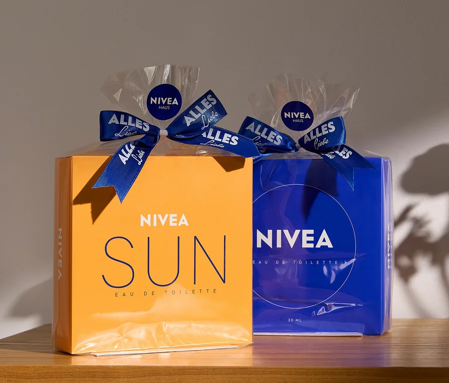 Zwei NIVEA Eau de Toilette Geschenksets auf einer Holzoberfläche: links ein orangefarbenes NIVEA SUN Set, rechts ein blaues NIVEA Set. Beide sind in Klarsichtfolie verpackt und mit einem blauen Band mit der Aufschrift "ALLES Liebe" und "NIVEA HAUS" verziert.