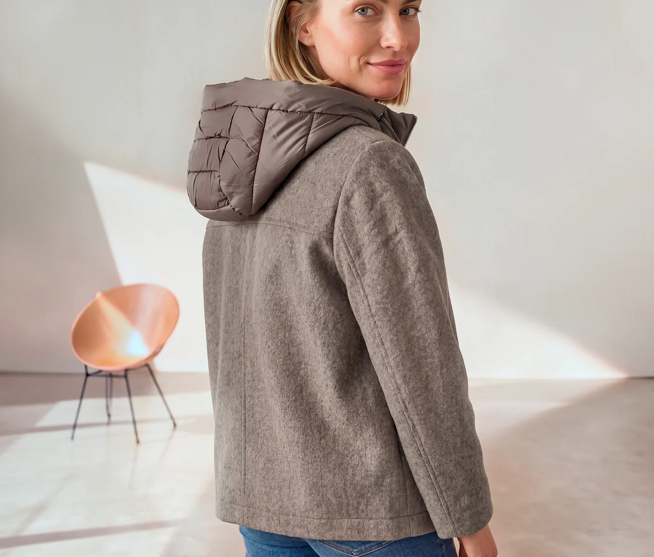 Eine Frau blickt über ihre Schulter in die Kamera, sie trägt eine graubraune Street One Übergangsjacke mit gesteppter Kapuze und blaue Jeans.