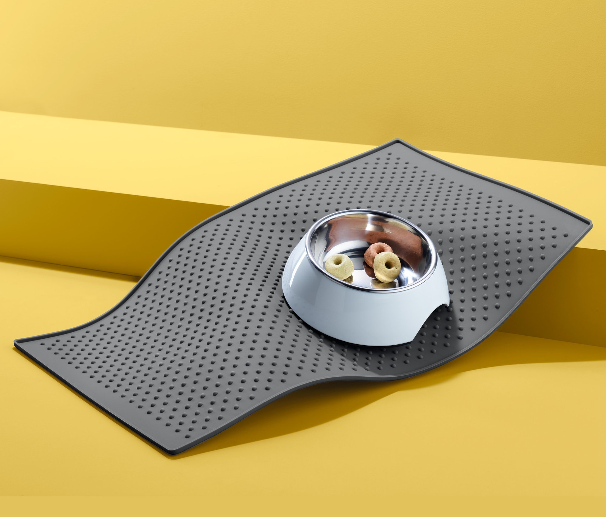 Grauer Futternapfuntersetzer mit einem mit Futter gefüllten Tiernapf auf gelbem Hintergrund.