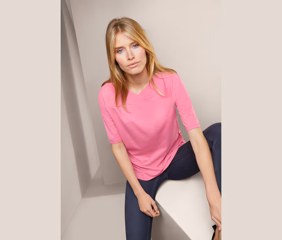Blonde Frau sitzt und trägt ein rosa Halbarmshirt.