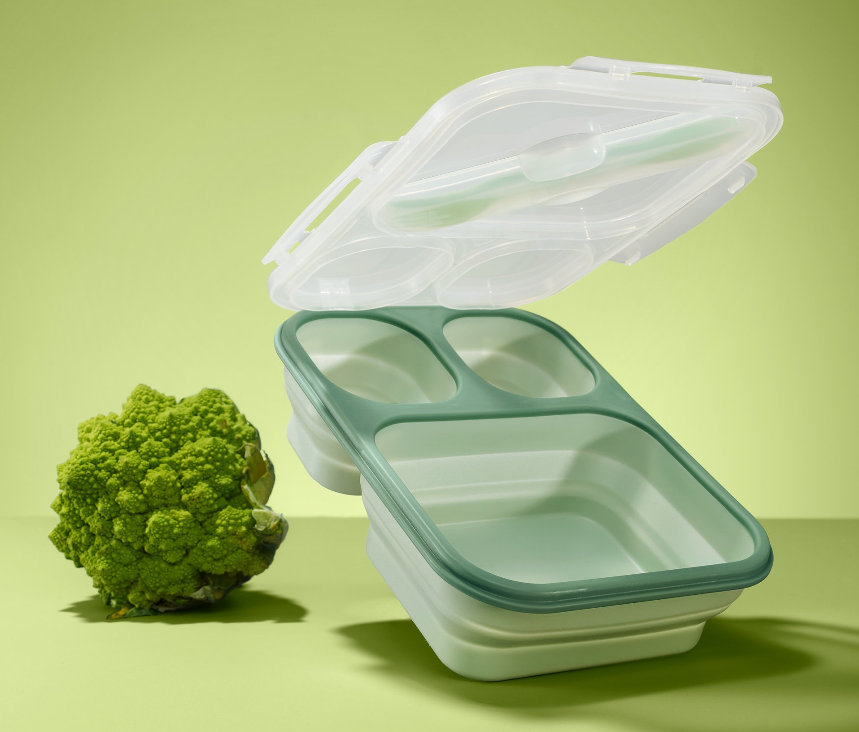 Eine grüne, faltbare Lunchbox mit drei Fächern und einem transparenten Deckel schwebt über einem Brokkoli auf grünem Hintergrund.