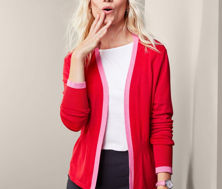 Blonde Frau schaut nachdenklich und trägt eine offene rote Feinstrickjacke.