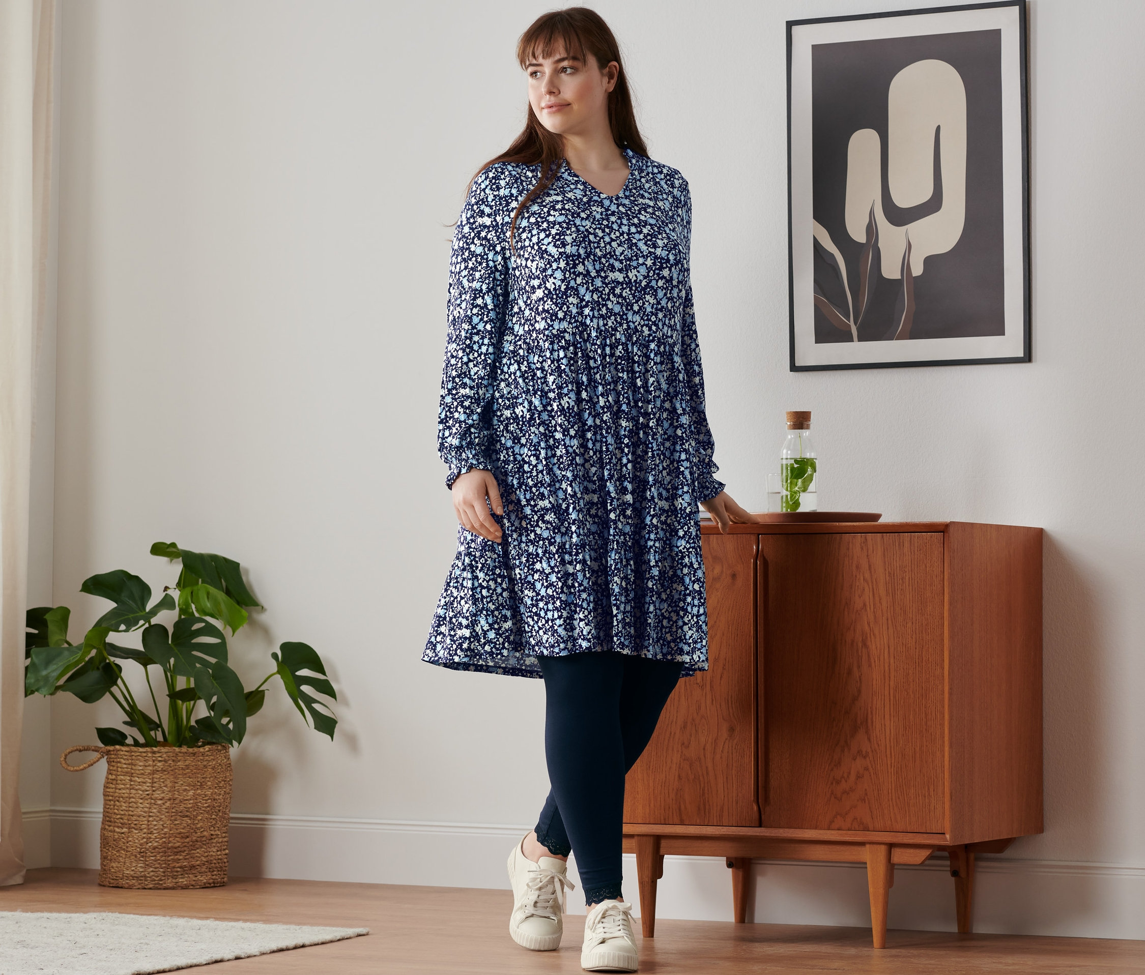 Eine Frau steht an einem Schrank und trägt ein florales Jerseykleid, Leggings mit Spitzenborte und Leder-Sneaker.