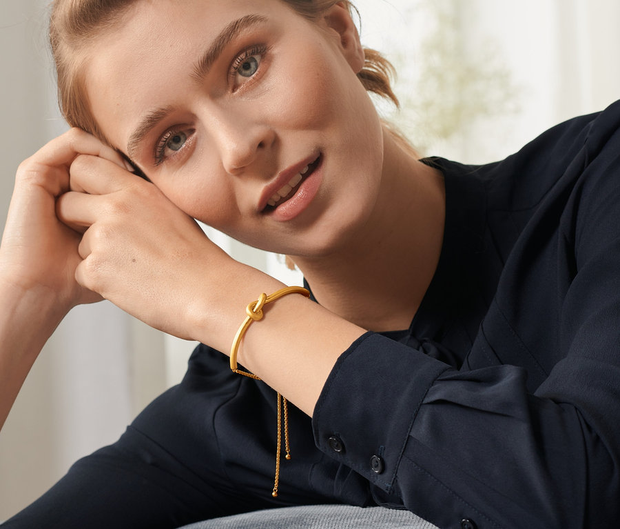 Frau mit blonden Haaren stützt ihren Kopf auf ihren Händen und trägt einen Slider-Armreif »Knot«, 18 Karat vergoldet, am Handgelenk.