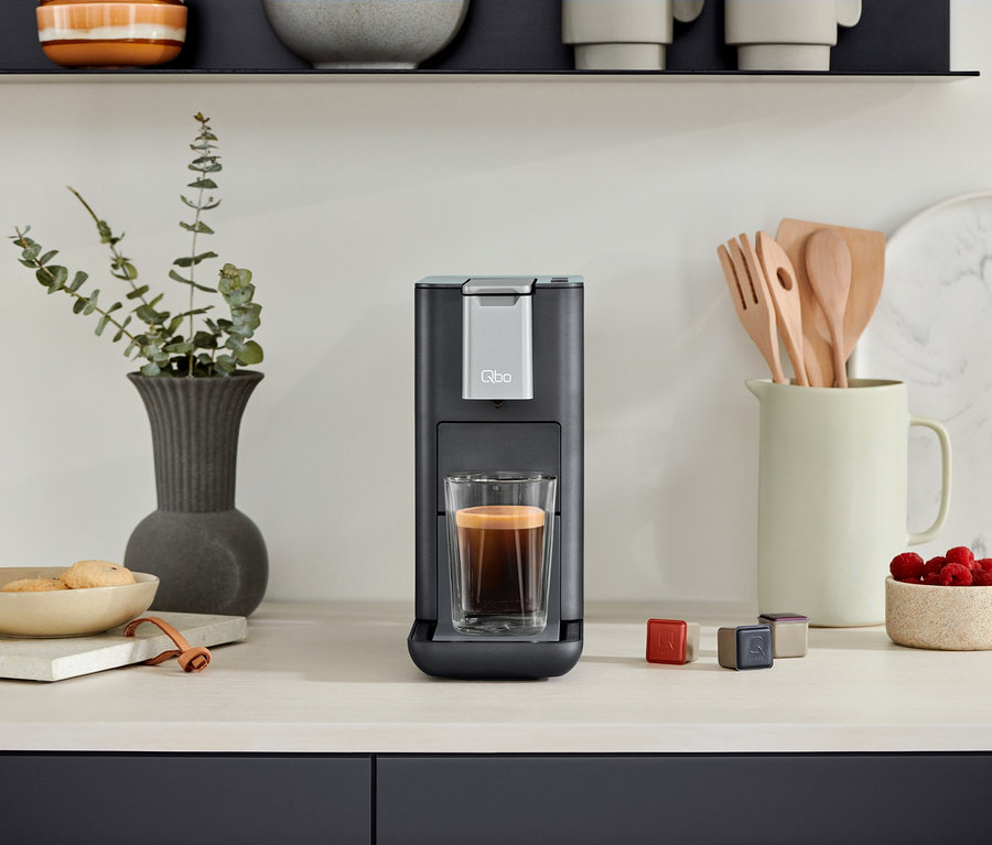 Eine Qbo ESSENTIAL Dark Stone Kaffeemaschine mit einer Tasse Kaffee. Daneben stehen Kapseln und eine Schale mit Himbeeren.