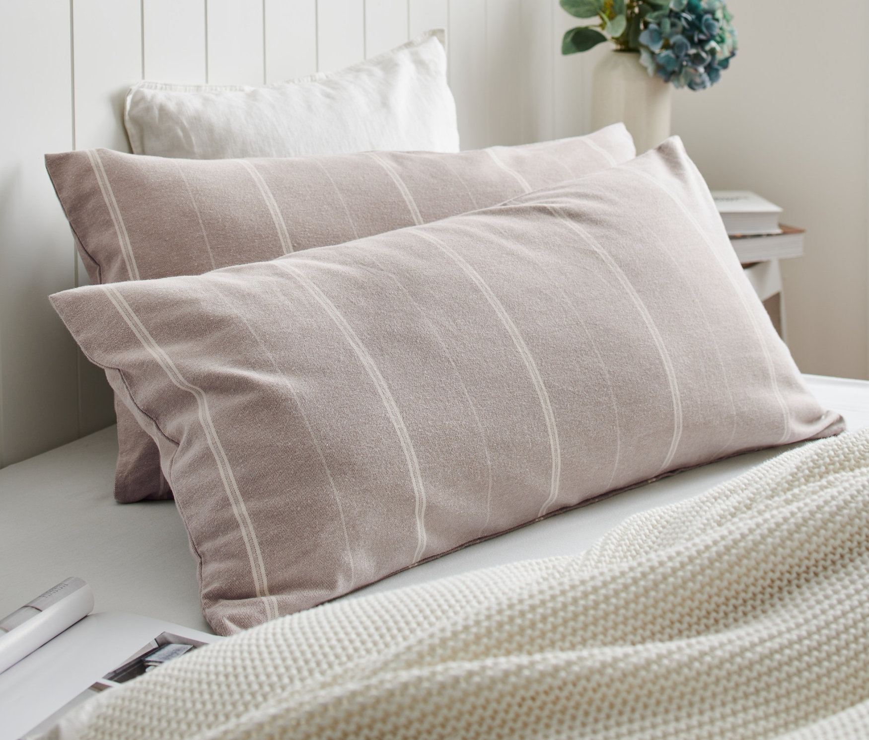 Zwei Edelflanell-Kopfkissenbezüge mit Streifen liegen auf einem Bett neben einer weißen Strickdecke.