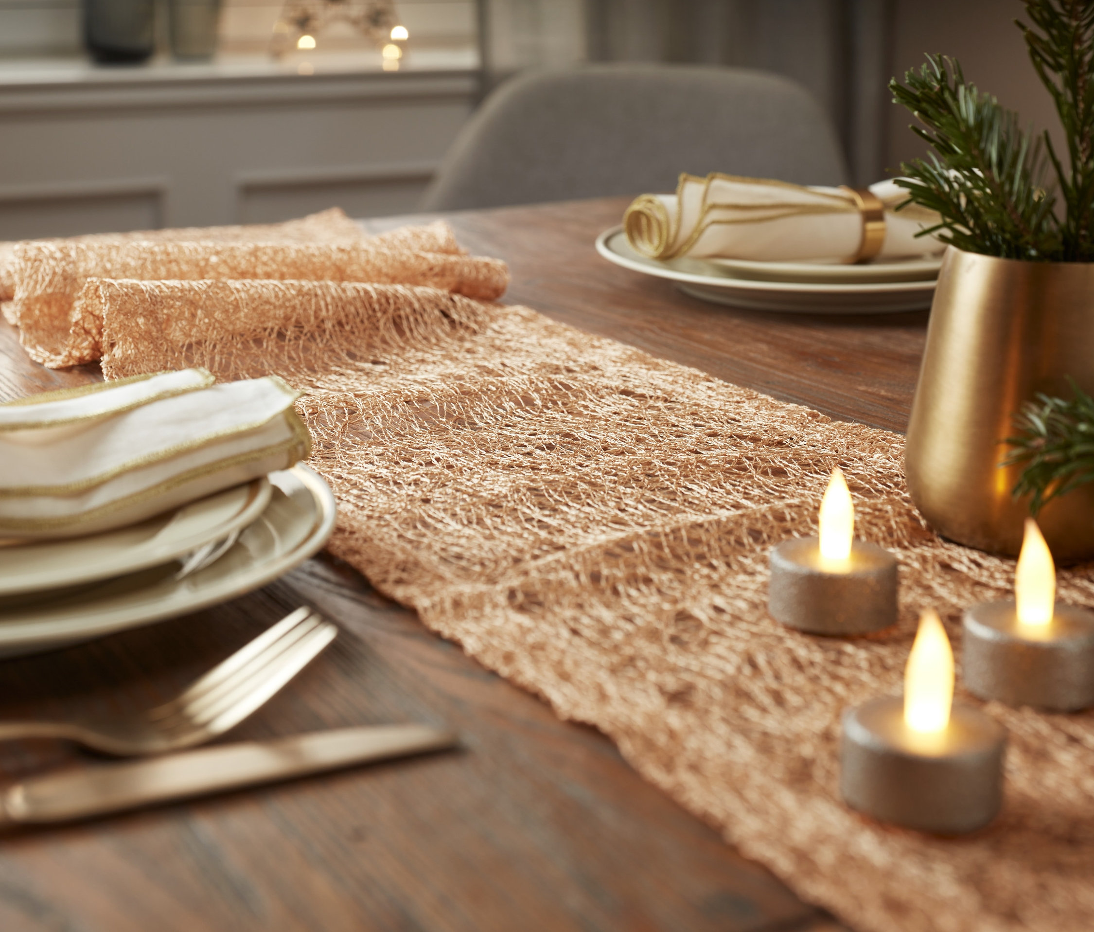 Auf einem Holztisch liegen ein beiger Tischläufer, drei brennende Kerzen und eine Vase mit Zweigen.