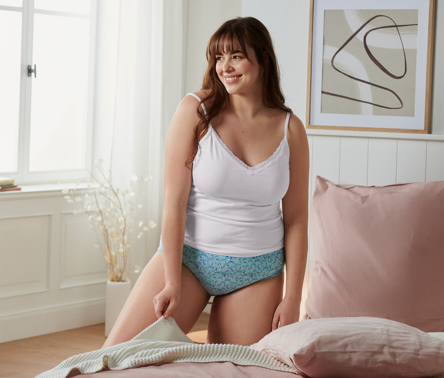 Frau kniet auf einem Bett und hält eine Decke. Sie trägt ein weisses Hemdchen und Pantys.