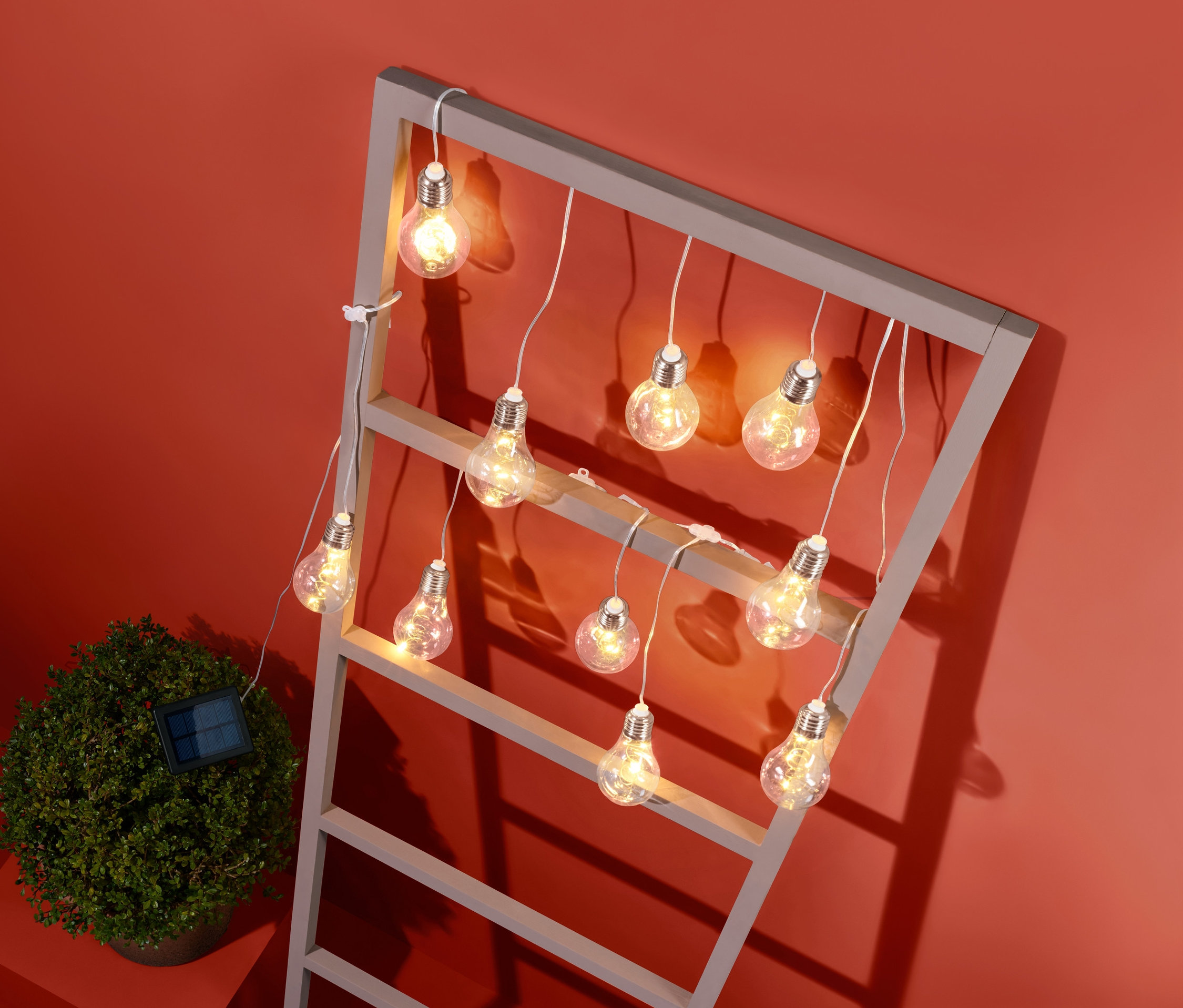 Eine Solar-Lichterkette hängt an einem grauen Rahmen, neben einem kleinen Busch vor einem orangefarbenen Hintergrund.