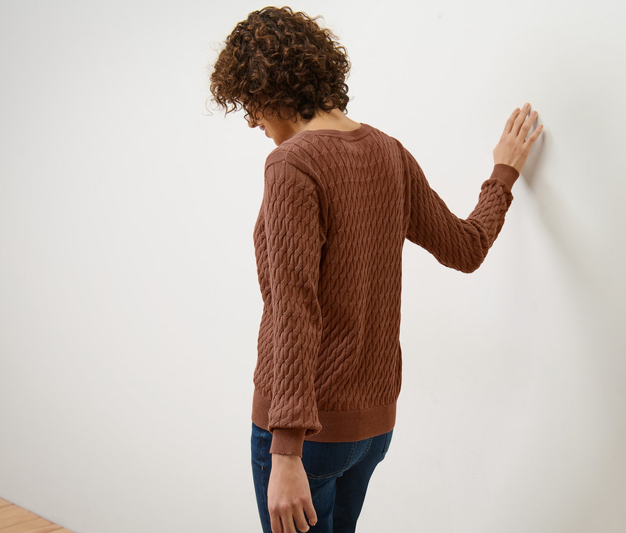 Frau lehnt an einer weißen Wand und trägt einen Zopfstrickpullover in Cognac.
