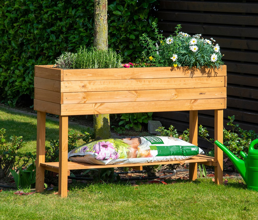 Holz-Hochbeet, ca. 117 x 80 x 40 cm, bepflanzt mit Kräutern und Blumen, auf einer Wiese mit einer grünen Gießkanne.