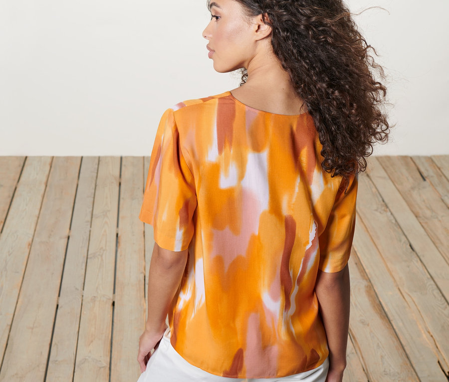 Frau von hinten, trägt eine Bluse mit Aquarell-Print und weiße Hose.