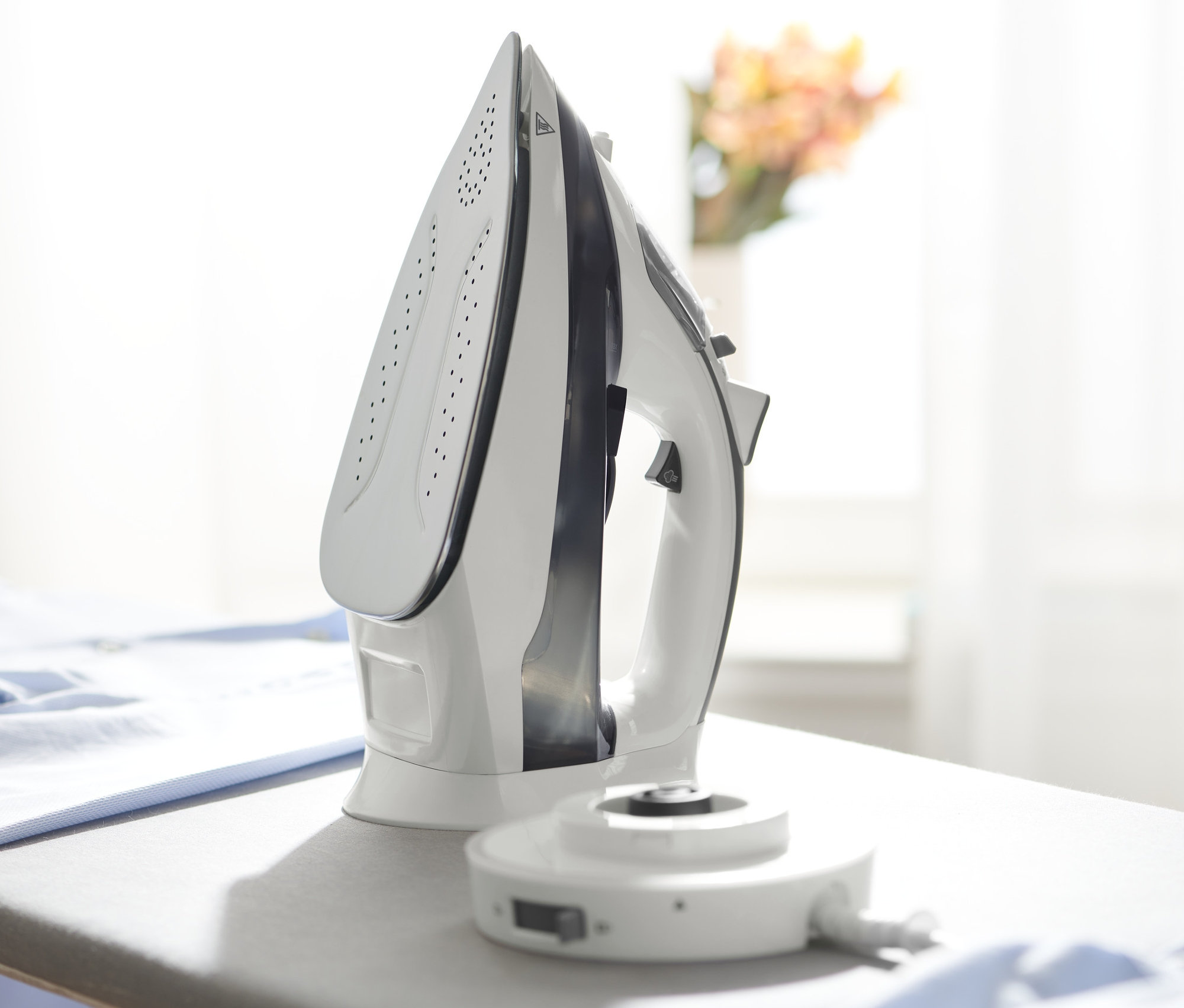 Ein weißes, kabelloses Dampfbügeleisen steht auf einem grauen Bügelbrett neben einem Haufen blauer Kleidung.