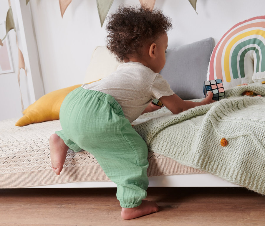 Kleinkind krabbelt auf ein Bett und spielt mit einem Zauberwürfel. Es trägt eine Baby-Schlupfwebhose.
