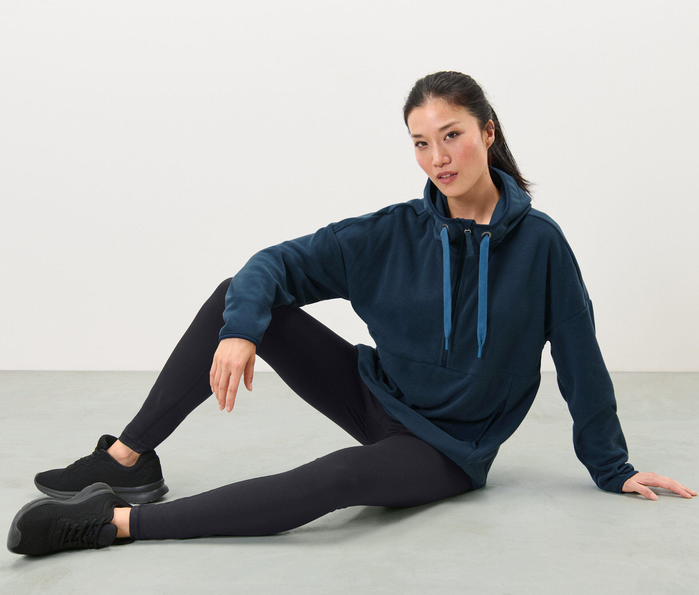 Frau sitzt am Boden und trägt einen Kapuzen-Fleece-Pullover, schwarze Leggings und Turnschuhe.