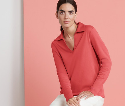 Frau sitzt auf einem durchsichtigen Hocker und trägt ein lachsfarbenes Sweatshirt mit Polokragen und eine weiße Hose.