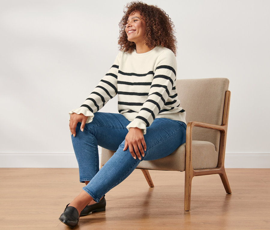 Frau sitzt in einem Sessel und trägt eine Slimfit-Jeans und einen gestreiften Strickpullover mit Rüschendetail.