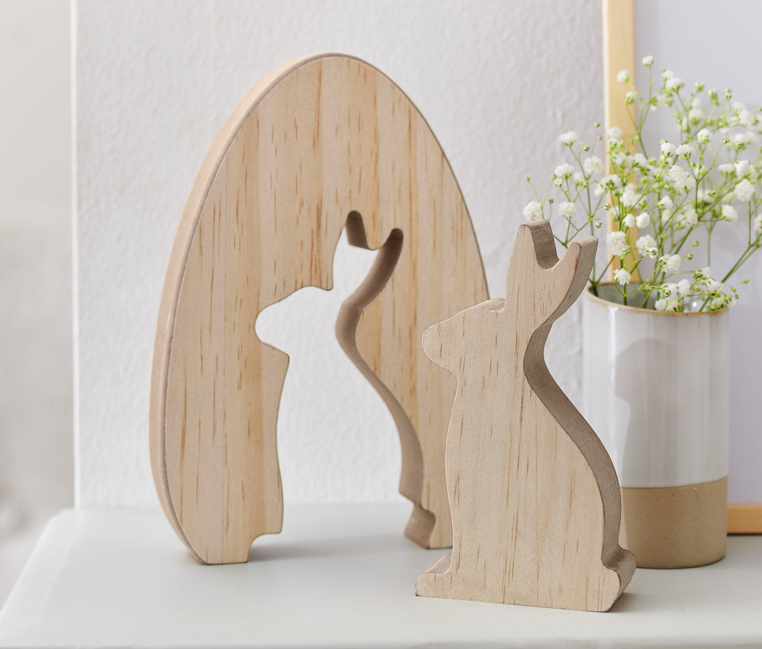 Zwei Deko-Hasen aus Holz stehen nebeneinander auf einem weißen Regal, dahinter eine Vase mit weißen Blumen.