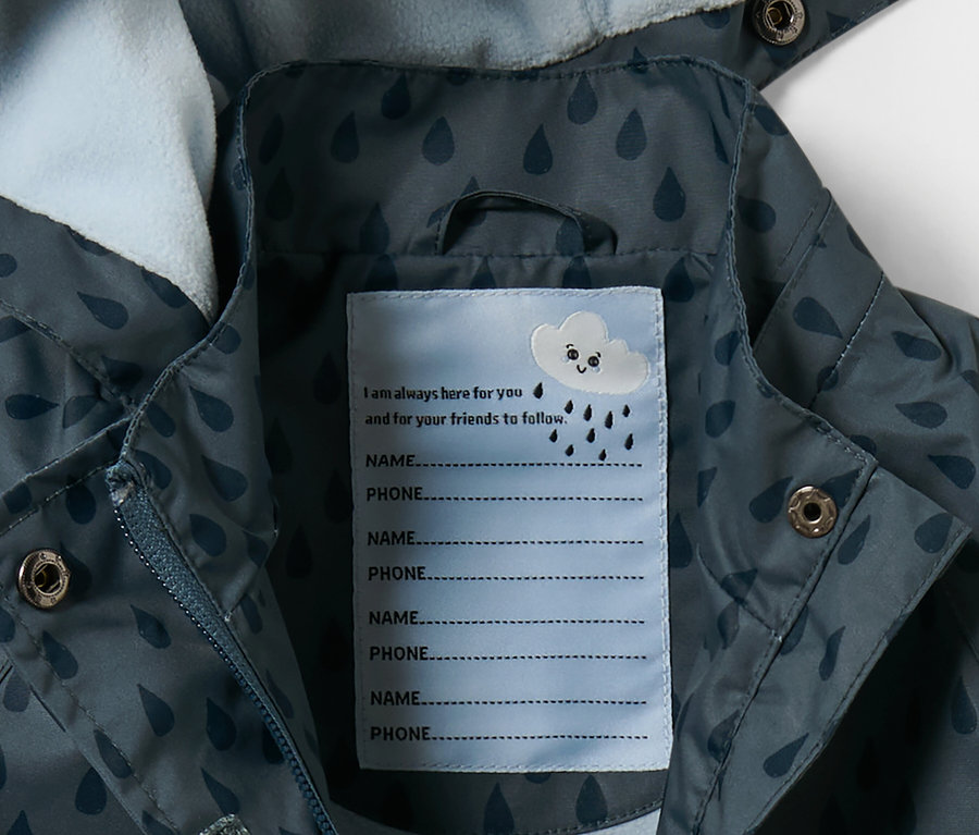 Detail einer Kleinkind-Thermo-Regenjacke, schieferblau Tropfen, mit einem Etikett für Name und Telefonnummer.